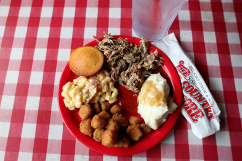 A red plate with pulled pork, cornbread, macaroni and cheese, mashed potatoes, fried okra, and a glass of water.