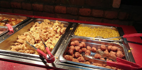 A buffet spread featuring fried shrimp, corn, and round brown snacks, with serving tongs on a red table.