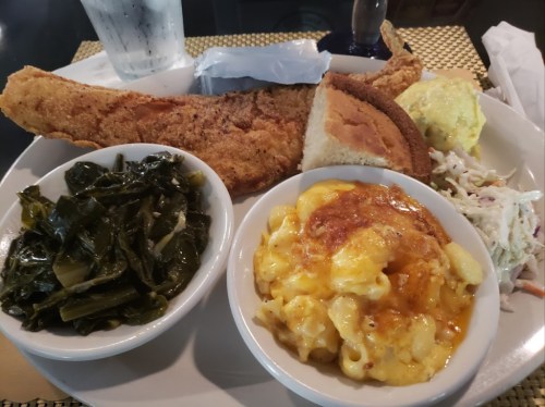 A plate of fried fish, cornbread, collard greens, macaroni and cheese, and coleslaw, served with a glass of water.