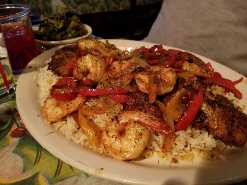 A plate of seasoned shrimp and fish over rice, topped with colorful bell peppers, served with a side of greens.
