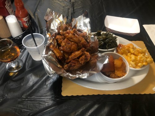 A plate of crispy chicken wings, collard greens, macaroni and cheese, and a drink on a black tablecloth.