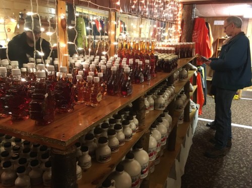 A man examines bottles of maple syrup on a wooden shelf, surrounded by festive lights in a cozy shop.