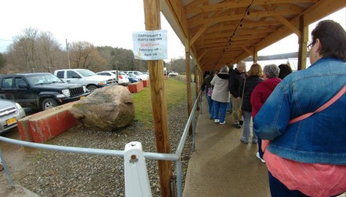 A line of people waiting under a covered walkway beside parked cars and a large rock, with a sign for Cartwright's Maple Tree Inn.