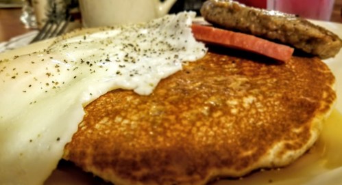 A close-up of a breakfast plate featuring a pancake, fried egg, sausage patty, and a slice of tomato.