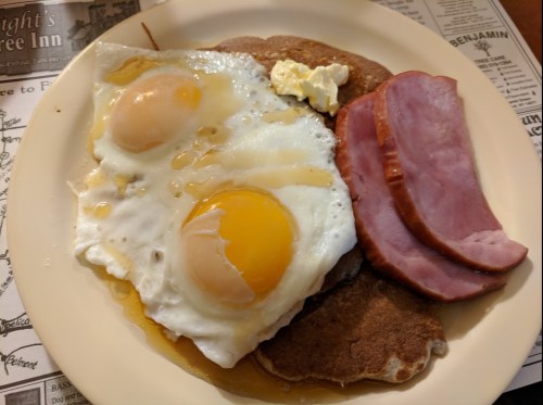 A plate with two fried eggs, ham slices, and a dollop of butter on a stack of pancakes.