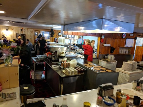 A busy diner kitchen with staff cooking pancakes, preparing food, and serving customers in a cozy, wooden interior.
