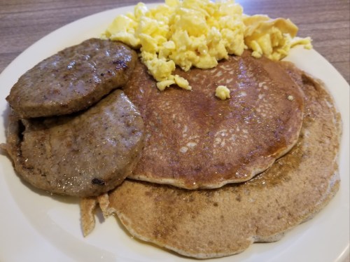 A plate of pancakes with two sausage patties and scrambled eggs, drizzled with syrup.