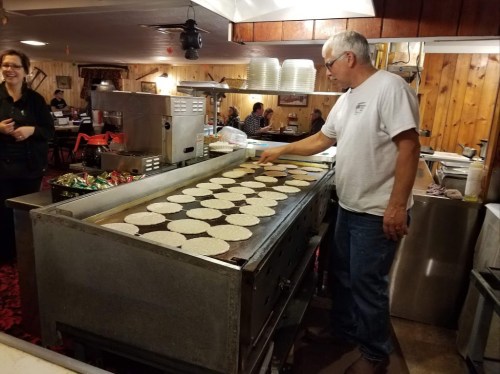 A man cooks multiple pancakes on a large griddle in a busy restaurant, with customers in the background.
