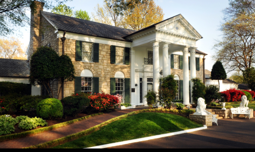 A large stone house with white columns, surrounded by lush greenery and colorful flowers in a well-maintained garden.