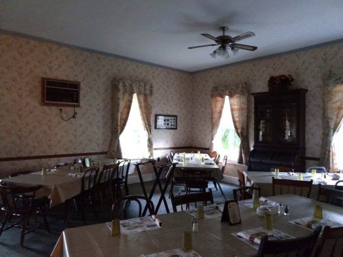 A dimly lit dining room with tables set for a meal, floral wallpaper, and a vintage piano in the corner.