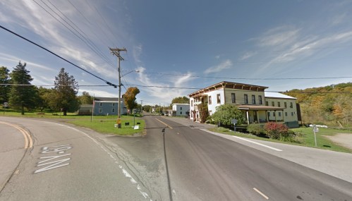 A small town intersection with a few buildings, trees, and a clear blue sky. Road signs are visible.