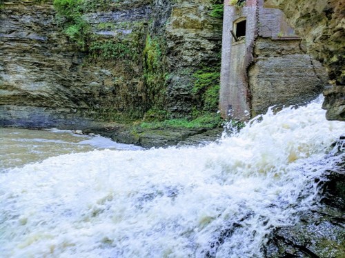 A rushing waterfall cascades over rocks near a moss-covered cliff and an old brick structure.