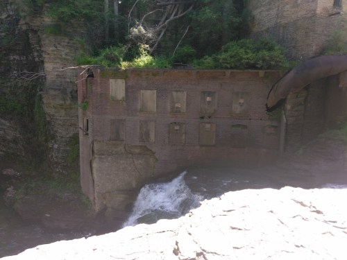 Abandoned brick building with boarded windows beside a flowing waterfall and lush greenery.