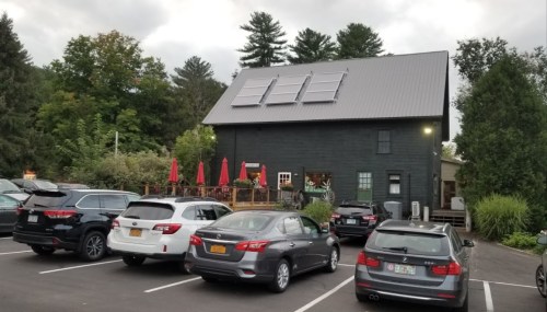 A black building with solar panels, surrounded by trees, and parked cars in front, under a cloudy sky.