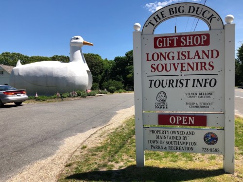 A large white duck statue next to a sign for a gift shop and tourist information on Long Island.