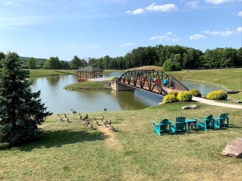 A scenic park with a pond, a bridge, and people walking. Adirondack chairs and geese are in the foreground.