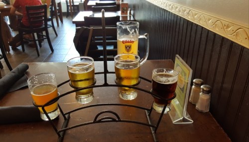 A flight of four small beer glasses on a metal holder, with a larger beer mug in the background on a restaurant table.