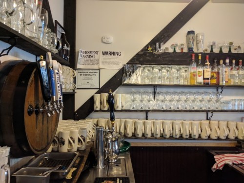 A bar with a wooden beer tap, shelves of glasses, and a variety of liquor bottles displayed on the wall.
