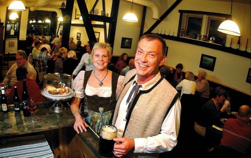 A smiling couple in traditional attire stands in a bustling restaurant, with guests dining in the background.