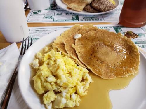 A plate with scrambled eggs and three pancakes drizzled with syrup, set on a table with menus in the background.