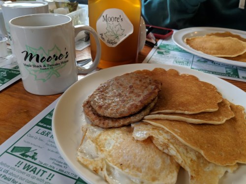 A plate with pancakes and sausage, alongside a coffee mug and maple syrup bottle from Moore's Maple Shack.