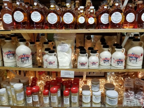 A display of various maple syrup bottles and containers on wooden shelves, with twinkling lights in the background.