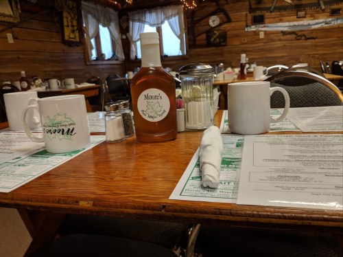 A cozy restaurant table set with menus, coffee mugs, a bottle of sauce, and condiments in a rustic wooden setting.