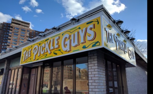 Sign for "The Pickle Guys" on a building, featuring colorful lettering against a bright blue sky.