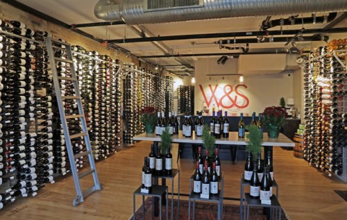 A wine shop interior featuring walls lined with wine bottles and a central display table with more bottles and plants.