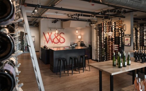 A modern wine shop interior featuring a bar, wine racks, and wooden tables with various wine bottles displayed.