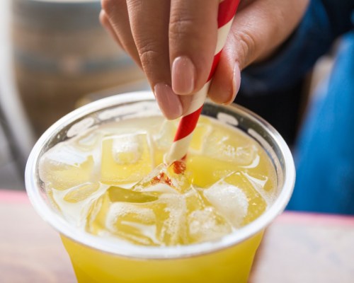 A hand with a striped straw stirs a refreshing yellow drink filled with ice in a clear cup.