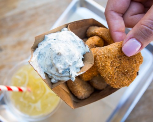 A hand holding a paper container of crispy nuggets with a side of creamy dipping sauce and a drink in the background.