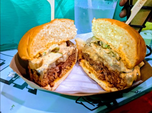 A close-up of a split burger with a thick patty, creamy sauce, and toppings, served in a paper tray.