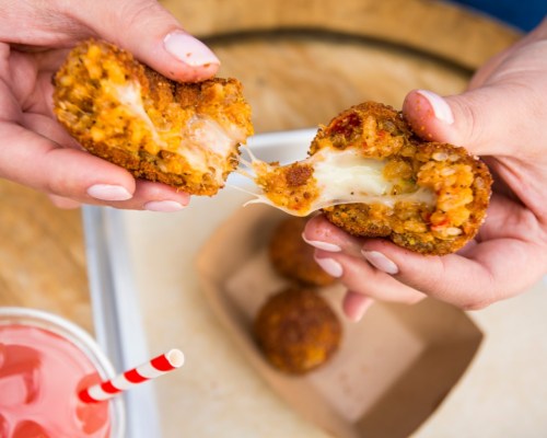 A person holds a crispy, cheese-filled ball, pulling it apart to reveal gooey cheese inside, with a drink nearby.