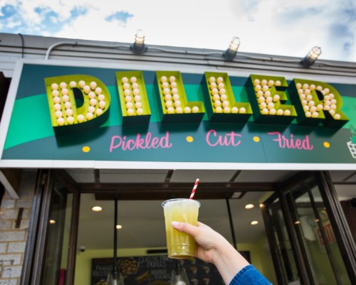 A hand holds a drink in front of a colorful sign that reads "DILLER" with lights, indicating a food establishment.
