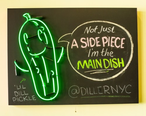 A neon sign featuring a cartoon pickle with the text: "Not just a side piece, I'm the main dish" @DILLERNYC.