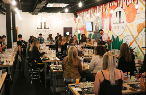 A lively bar setting with people seated at long tables, enjoying drinks and a presentation in a colorful, vibrant space.