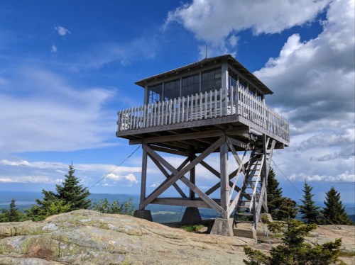 A wooden fire lookout tower on a rocky outcrop, surrounded by trees under a blue sky with scattered clouds.