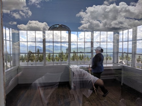 A person stands in a bright room with large windows, overlooking a scenic view of mountains and clouds.