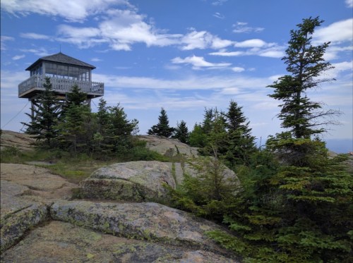 A lookout tower atop a rocky hill, surrounded by evergreen trees under a blue sky with scattered clouds.