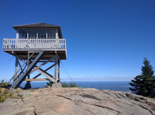 A wooden lookout tower on a rocky outcrop, surrounded by trees and a clear blue sky.