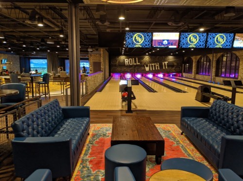 A modern bowling alley with vibrant lighting, plush seating, and a wooden table, creating a lively atmosphere.
