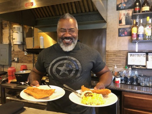 A smiling man in a gray shirt holds two plates of fried fish and scrambled eggs in a restaurant kitchen.
