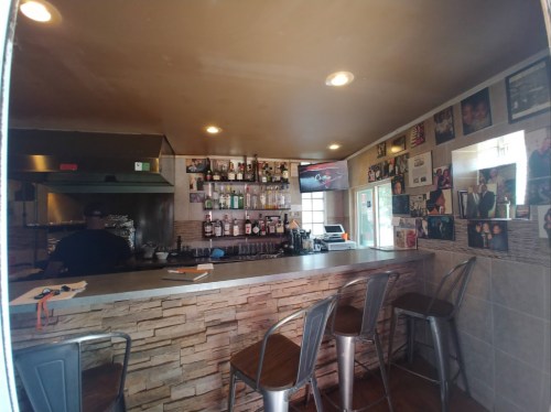 A cozy bar area with a counter, shelves of liquor, and photos on the walls, featuring a person in the background.