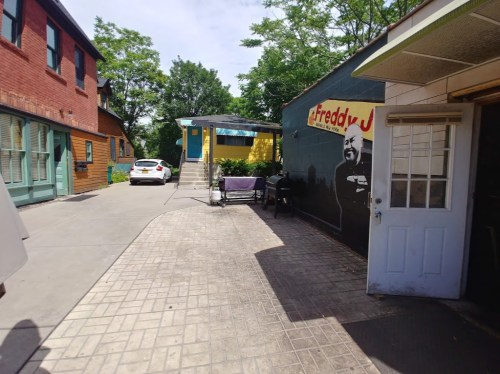 A pathway between buildings, featuring a mural of a man, a parked car, and outdoor seating.