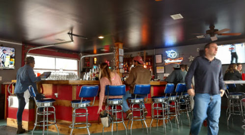 A lively bar scene with patrons at the counter, colorful decor, and a TV showing a sports game in the background.