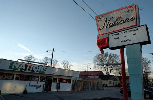 A retro sign for "The Nations" restaurant with a message about struggle and progress, alongside a colorful building.