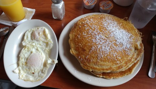 A plate of fluffy pancakes dusted with powdered sugar, alongside two sunny-side-up eggs and a glass of orange juice.