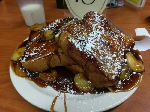 A plate of French toast topped with powdered sugar and caramelized bananas, drizzled with syrup.