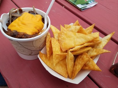A cup of chili topped with melted cheese next to a serving of crispy tortilla chips on a red table.